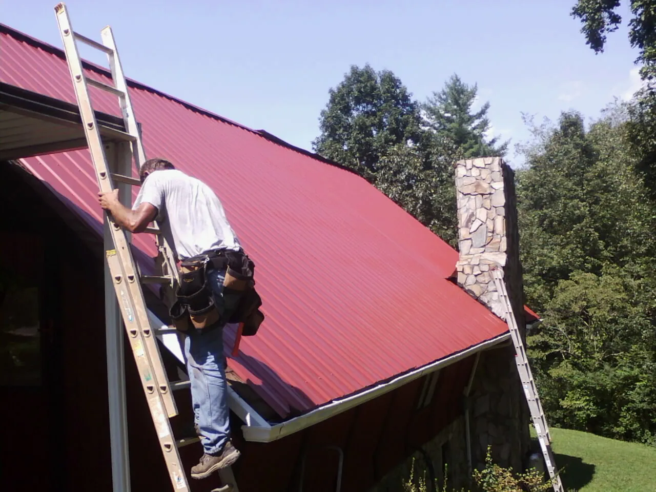 Roofer on a ladder beside a red metal roof and stone chimney — Soffit & Fascia Repair by Roofing Brothers in Hartville